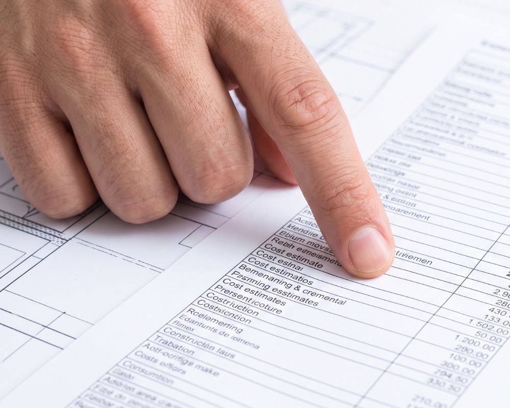 A detailed construction budget and cost estimate sheet are reviewed alongside a residential floor plan blueprint.