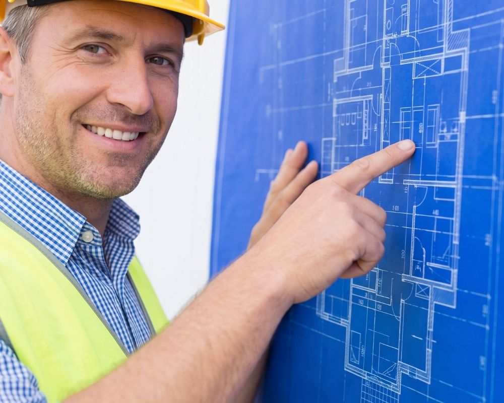 A construction project manager smiles as he points confidently to a specific detail on a set of complex building blueprints.